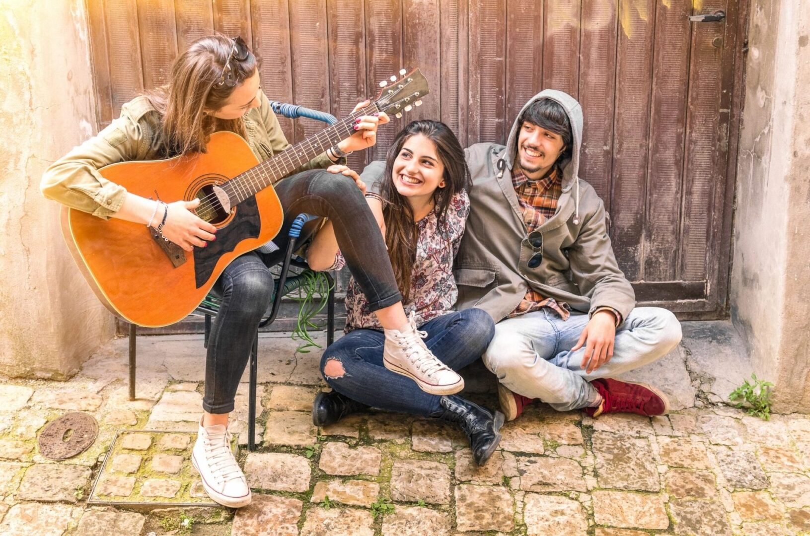 Three people sitting on a brick floor with one playing the guitar